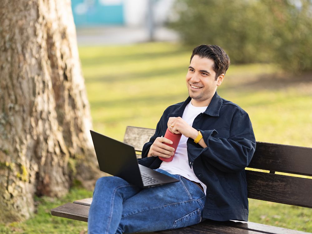 Een Henkel werknemer zit op een bank in een park en werkt op zijn laptop.