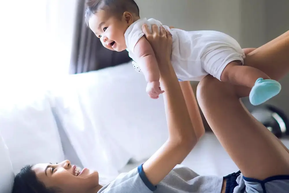 Een moeder tilt een lachende baby op en speelt ermee binnenshuis in een lichte huiselijke omgeving.