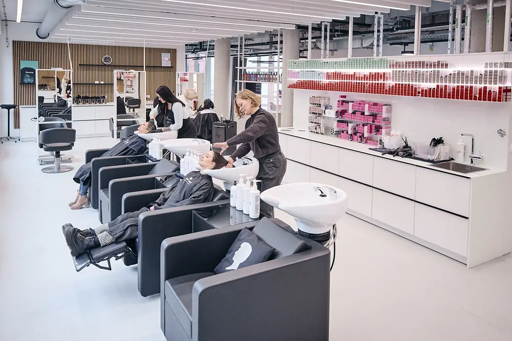 Ein Foto des modern eingerichteten Henkel-Testsalons in Hamburg, das drei Kundinnen in schwarzen Waschstühlen zeigt, die von Friseurinngen Haarbehandlungen erhalten. 