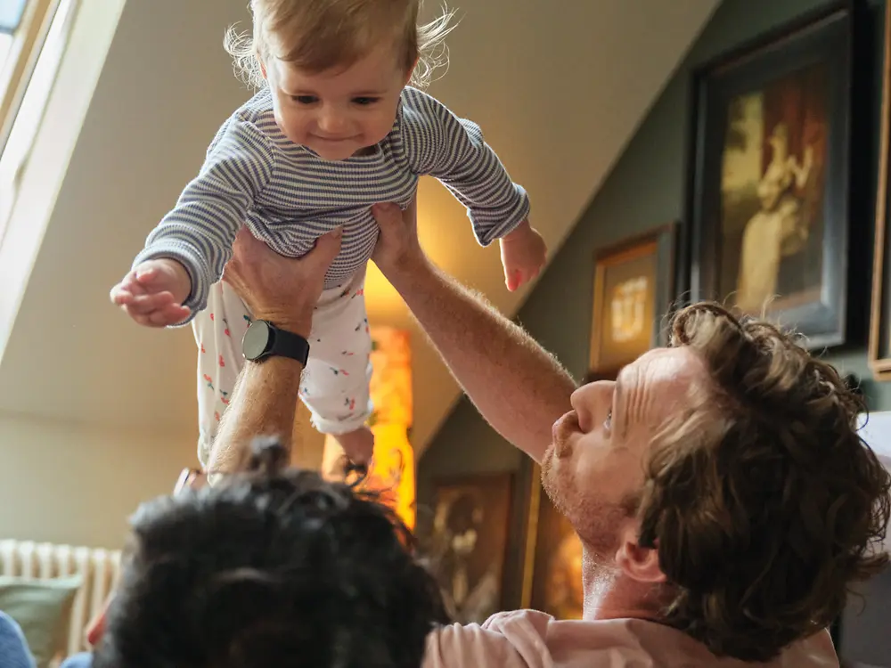 Twee vaders houden hun baby vast en glimlachen thuis, als symbool van inclusiviteit en Henkels gezinsvriendelijke ouderschapsverlofbeleid.