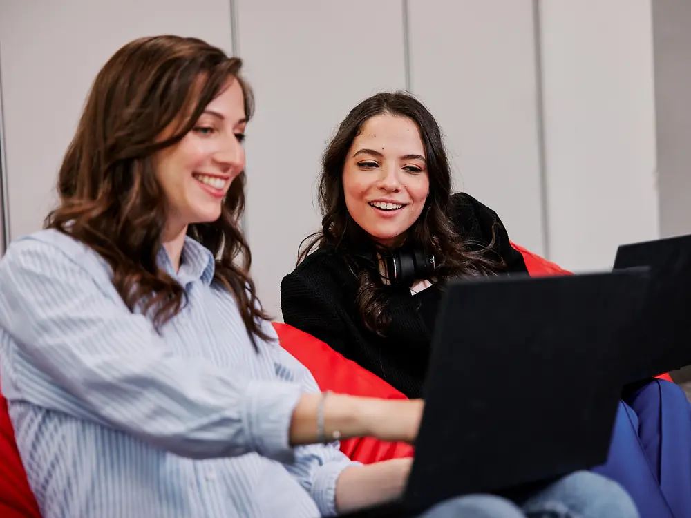 Twee mensen zitten op kleurrijke zitzakken in een moderne kantooromgeving, elk met een laptop. Ze lijken samen in gesprek te zijn over de personeelsvoordelen van Henkel, terwijl ze gezamenlijk aan het werk zijn.