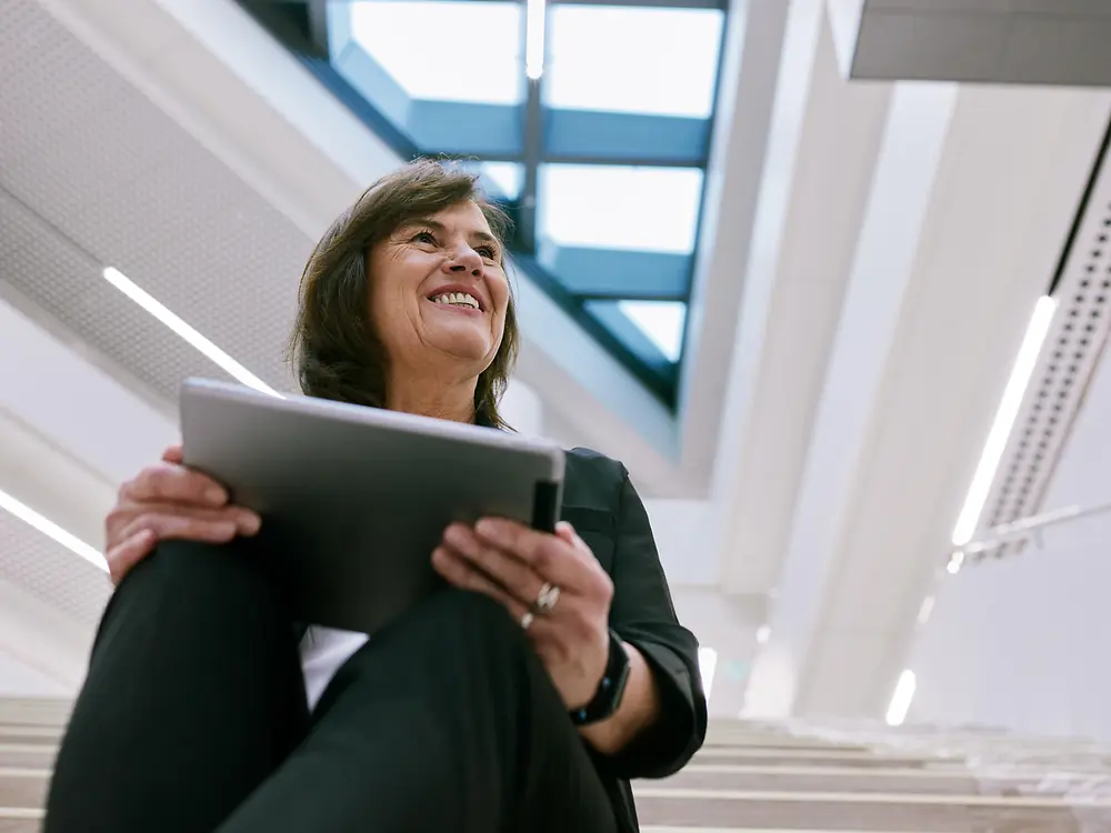Persoon die op een binnentrap in een modern gebouw zit, met een tablet. De achtergrond toont een helder, open architecturaal ontwerp met grote ramen en lichtkoepels, wat een nette en professionele sfeer creëert.