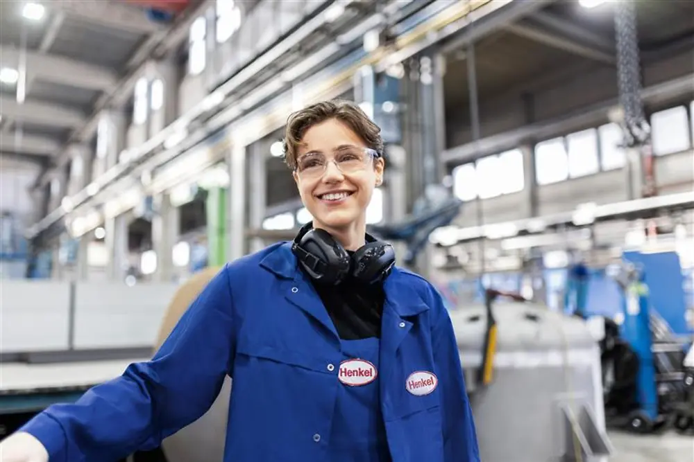 Glimlachende Henkel-medewerker in een blauwe werkjas en met gehoorbescherming, staand in een industriële productiehal, ter illustratie van expertise en innovatie in de productie.