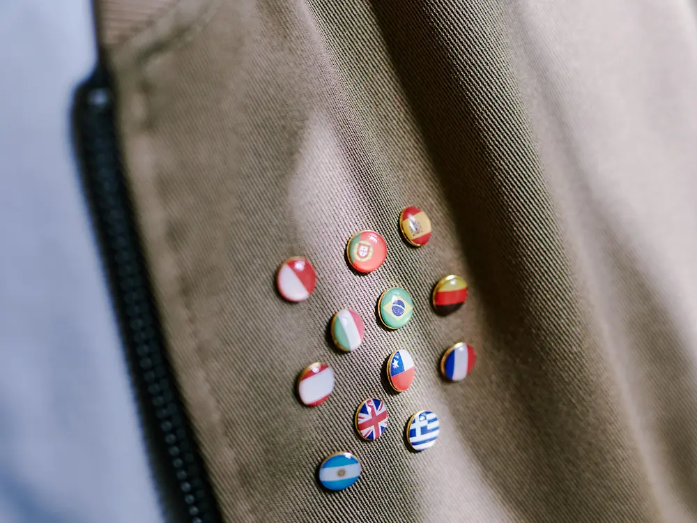 Close-up van een bruine stof met een rits aan de linkerkant en verschillende kleine ronde pins met diverse nationale vlaggen, als symbool van internationale diversiteit.