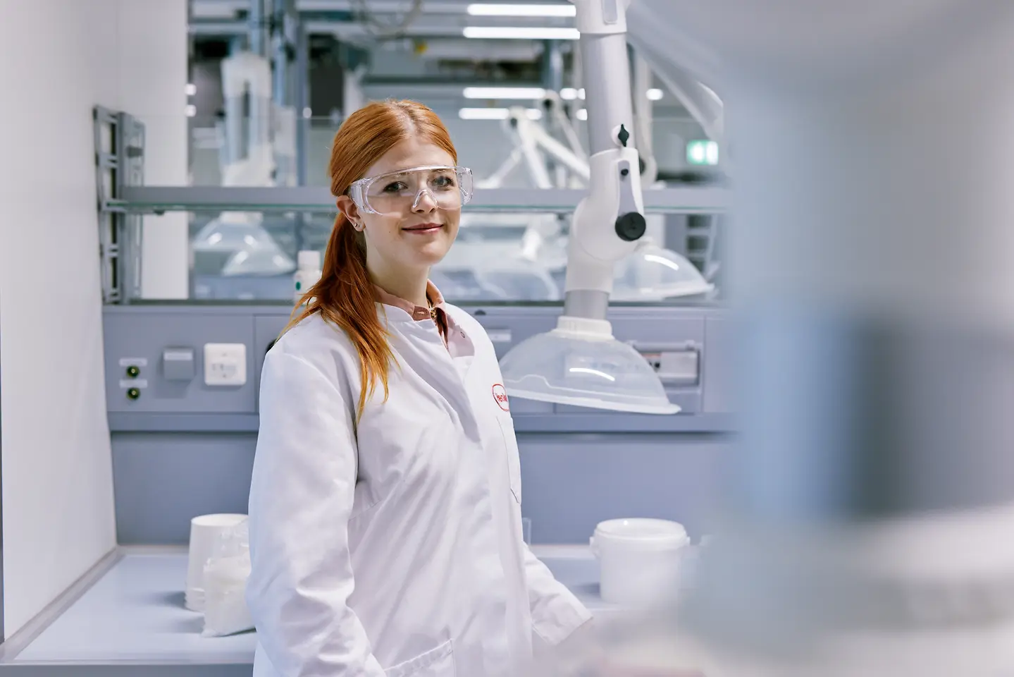 Pas afgestudeerde die zijn carrière start bij Henkel, met een witte laboratoriumjas, in een moderne laboratoriumomgeving met technische apparatuur op de achtergrond.