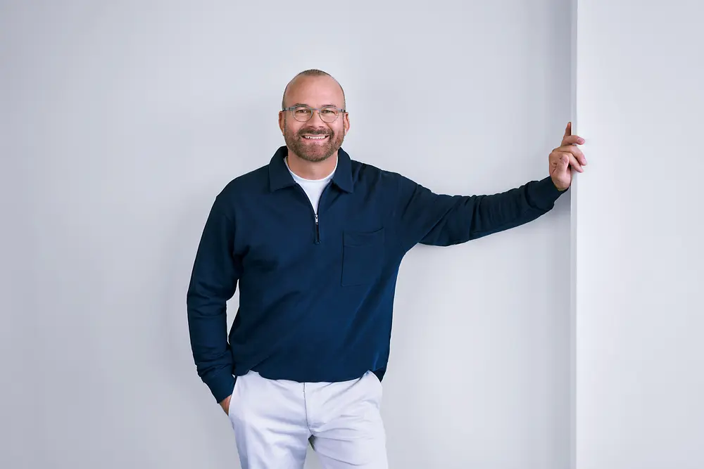 Ervaren professional bij Henkel, die zelfverzekerd tegen een witte muur staat in een moderne setting, met een donker shirt met lange mouwen en een lichtgekleurde broek.