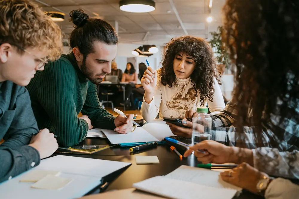 Een groep mensen aan een tafel in een moderne werkruimte binnen, terwijl ze samenwerken aan documenten, laptops en digitale toestellen. Verschillende pennen, papieren en post-its liggen over de tafel verspreid terwijl de groep discussieert en aantekeningen maakt.