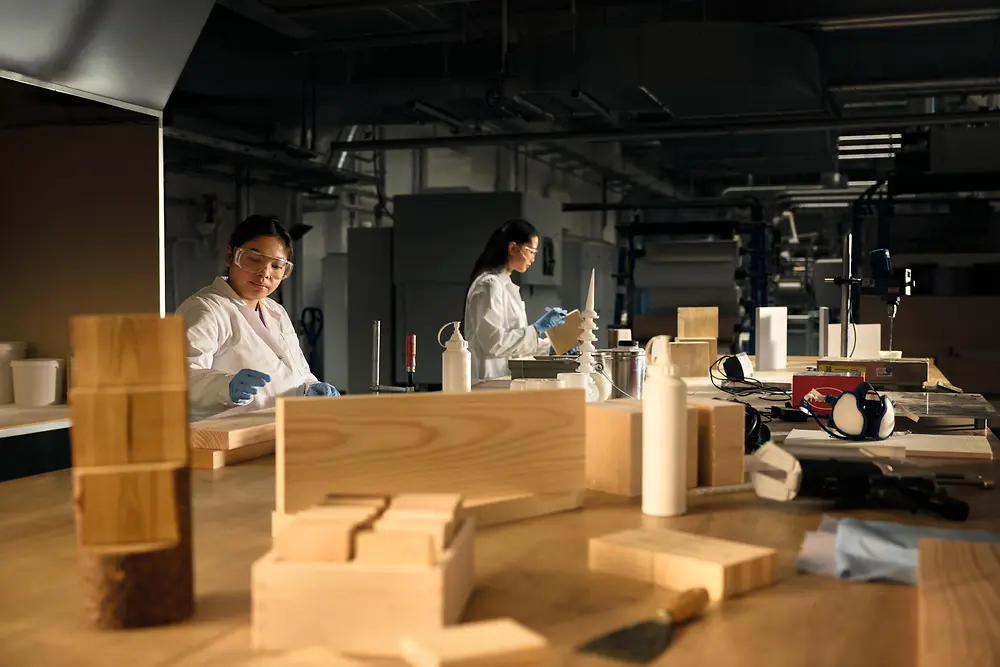 Workshop met personen in beschermende labojassen en handschoenen, aan het werk aan een grote houten tafel met houtstalen, gereedschap en flessen.