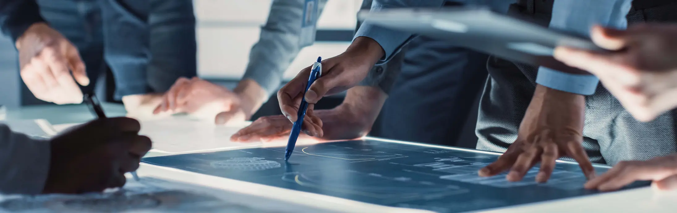 Close-up van verschillende mensen samen rond een grote, verlichte tafel, terwijl ze technische blauwdrukken bekijken en er aantekeningen bij maken met pennen en digitale toestellen.