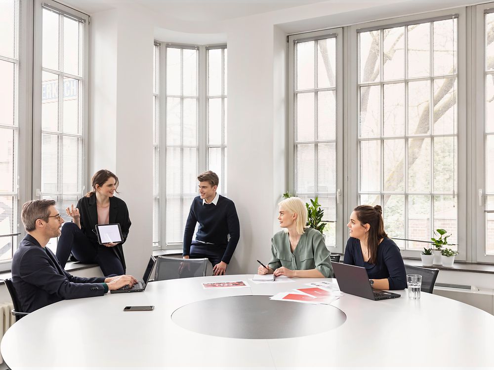5 mensen praten met elkaar zitten en staan rond een witte tafel in een Henkel-vergaderruimte met hoge ramen 